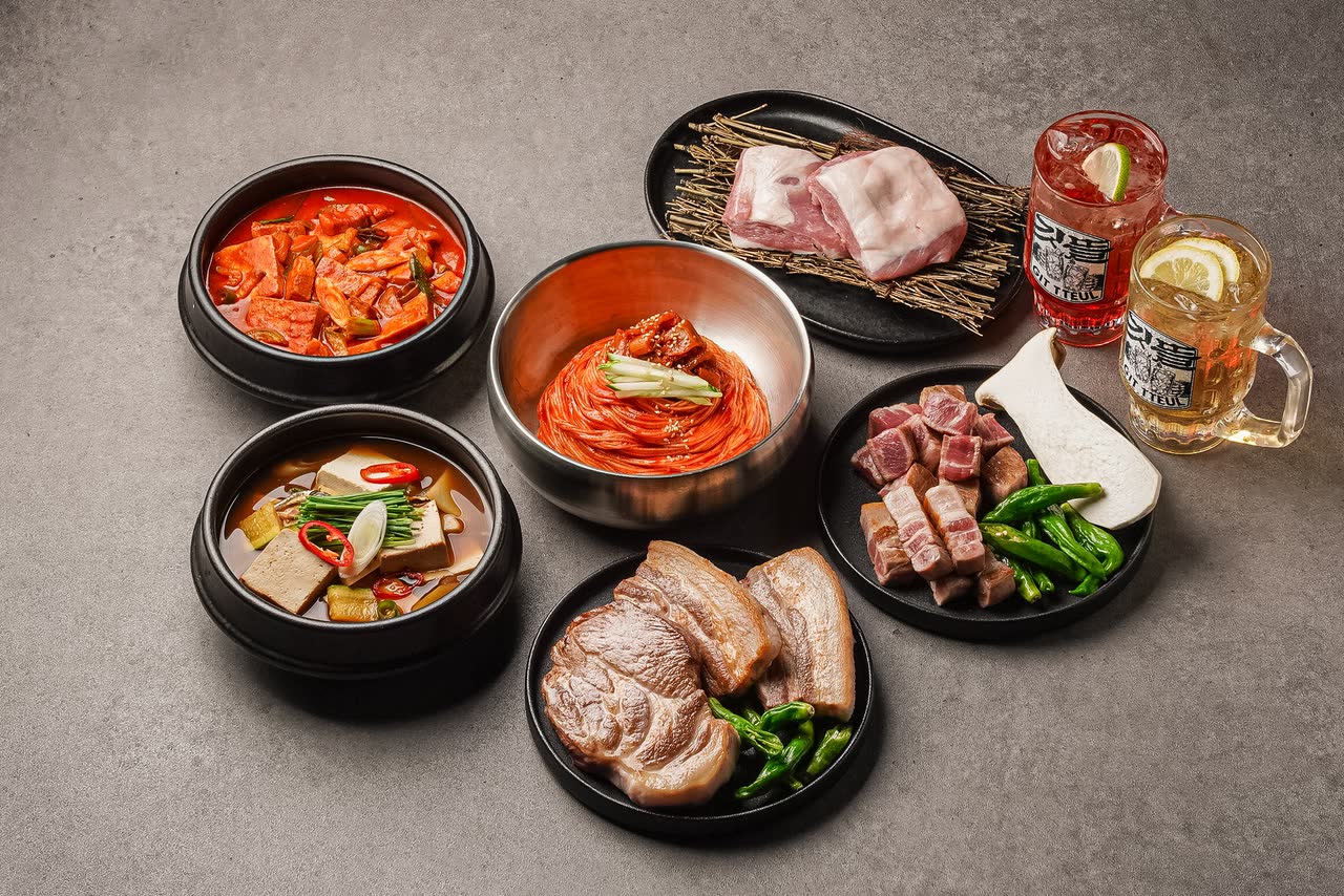 삼겹살과 목살을 담은 접시, 붉은 김치찌개, 나물 반찬 종지들, 그리고 레몬을 띄운 하이볼이 회색 상판 위에 둥글게 놓여 있다